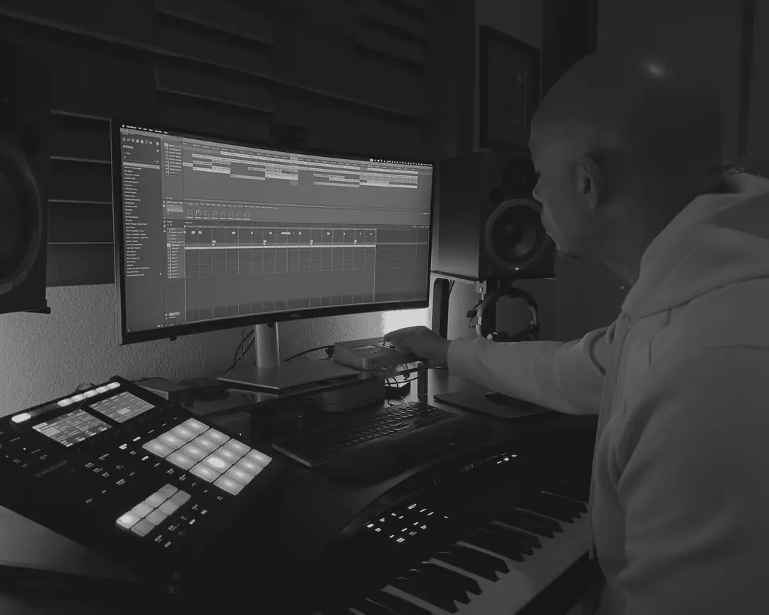 Moody at the desk reaching for the audio interface, Maschine pads lit and a session open on the monitor